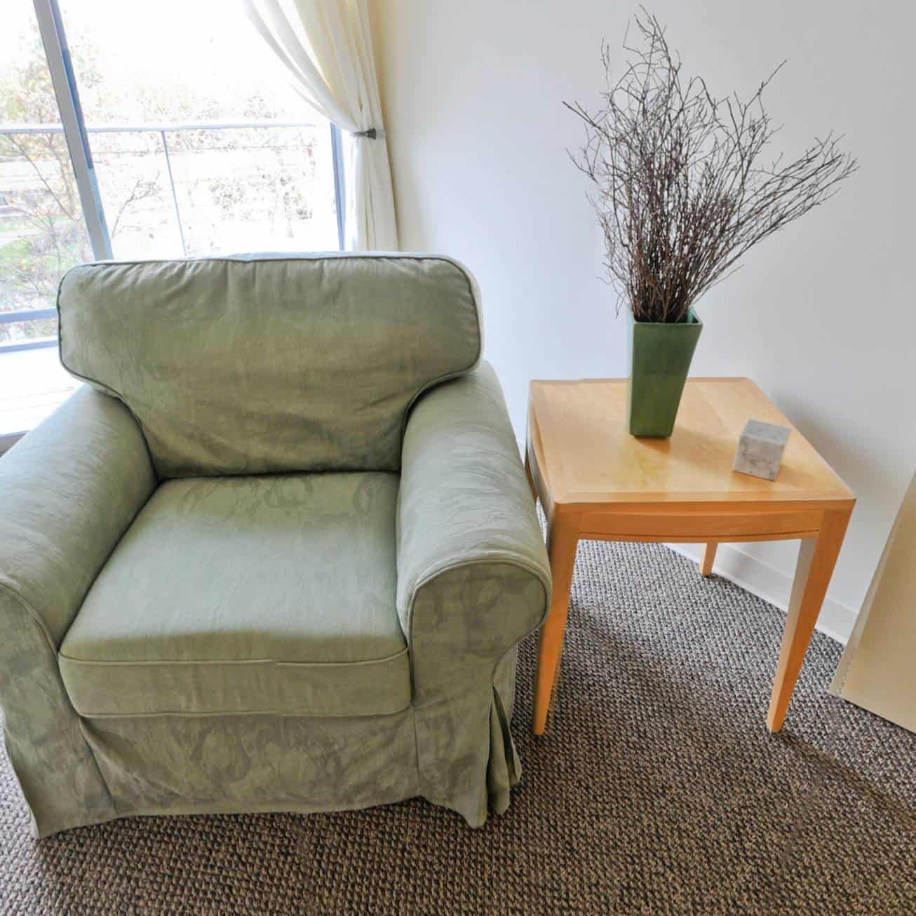A green upholstered armchair sits next to a small wood side table with a green vase containing branches and a small gray cube on a carpeted floor in front of a window.