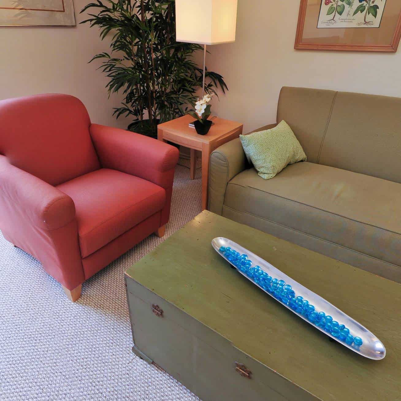 A living room with a red armchair, a green sofa, a wooden table with a glass bowl of blue marbles, a lamp, a small table with a plant, and a large plant in the corner. White walls and framed art.
