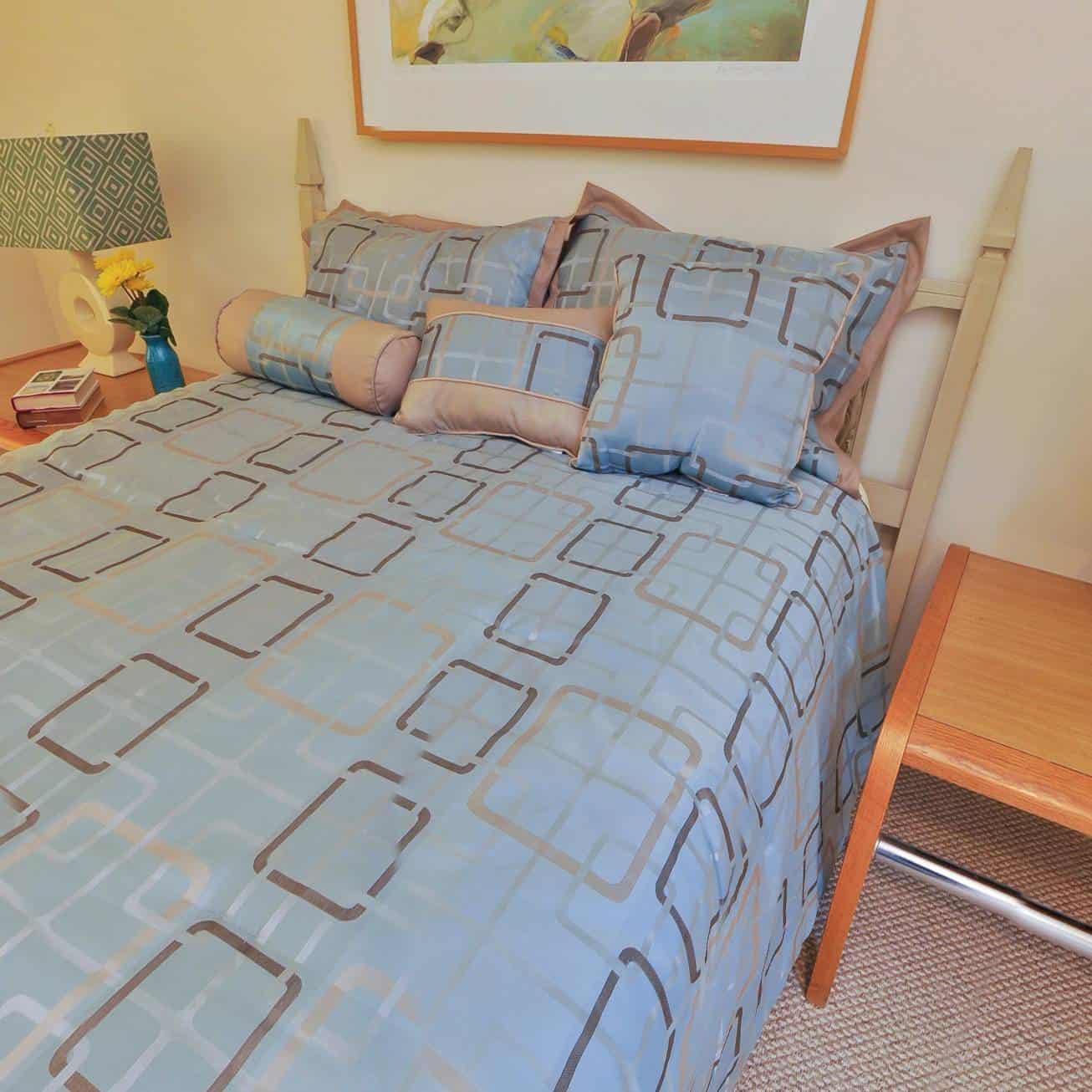 A neatly made bed with light blue and beige bedding featuring a geometric pattern, multiple pillows, and a wooden side table with a lamp, book, and vase of flowers.
