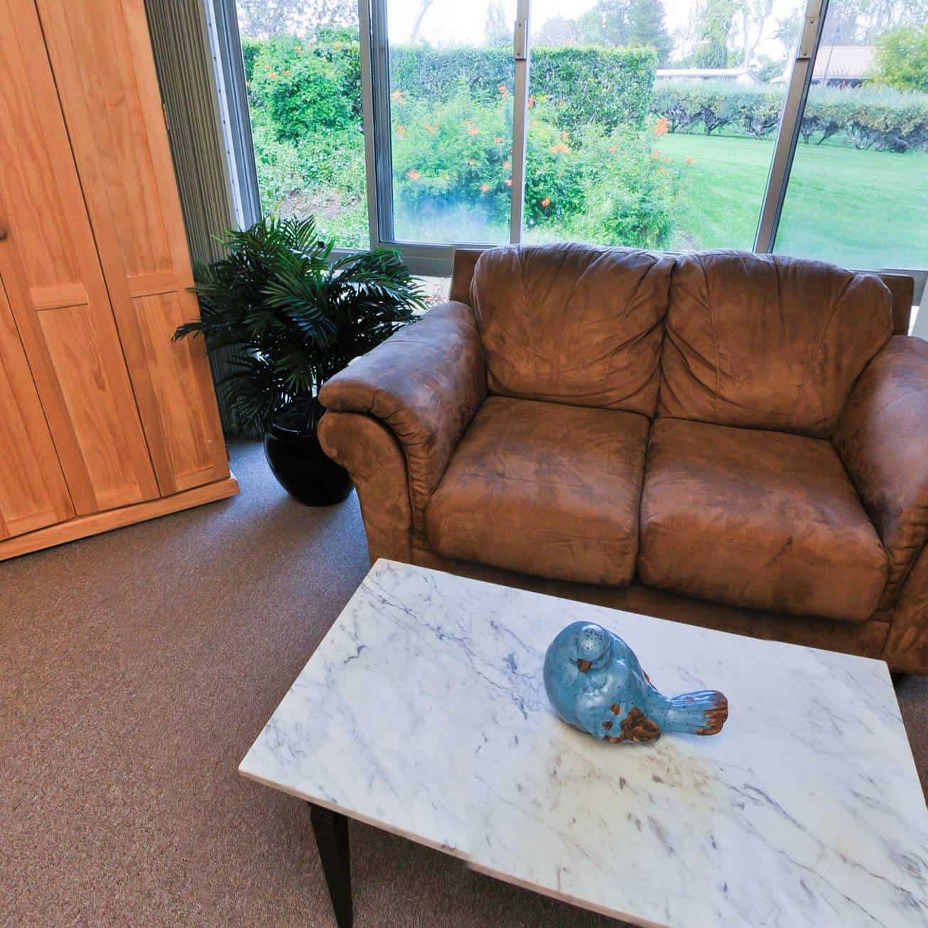 A brown leather sofa sits in front of a large window overlooking a garden. A white marble coffee table with a blue ceramic bird is in front of the sofa, and a wooden cabinet is on the left.