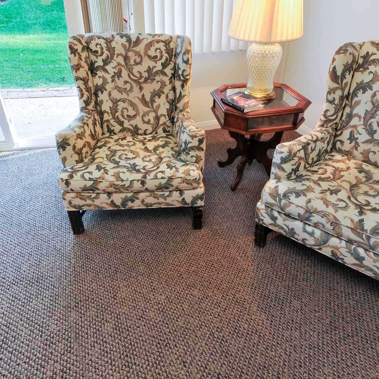 Two patterned armchairs are placed on a carpeted floor, with an octagonal wooden side table and a lamp between them. The room has vertical blinds and a sliding door leading outside.