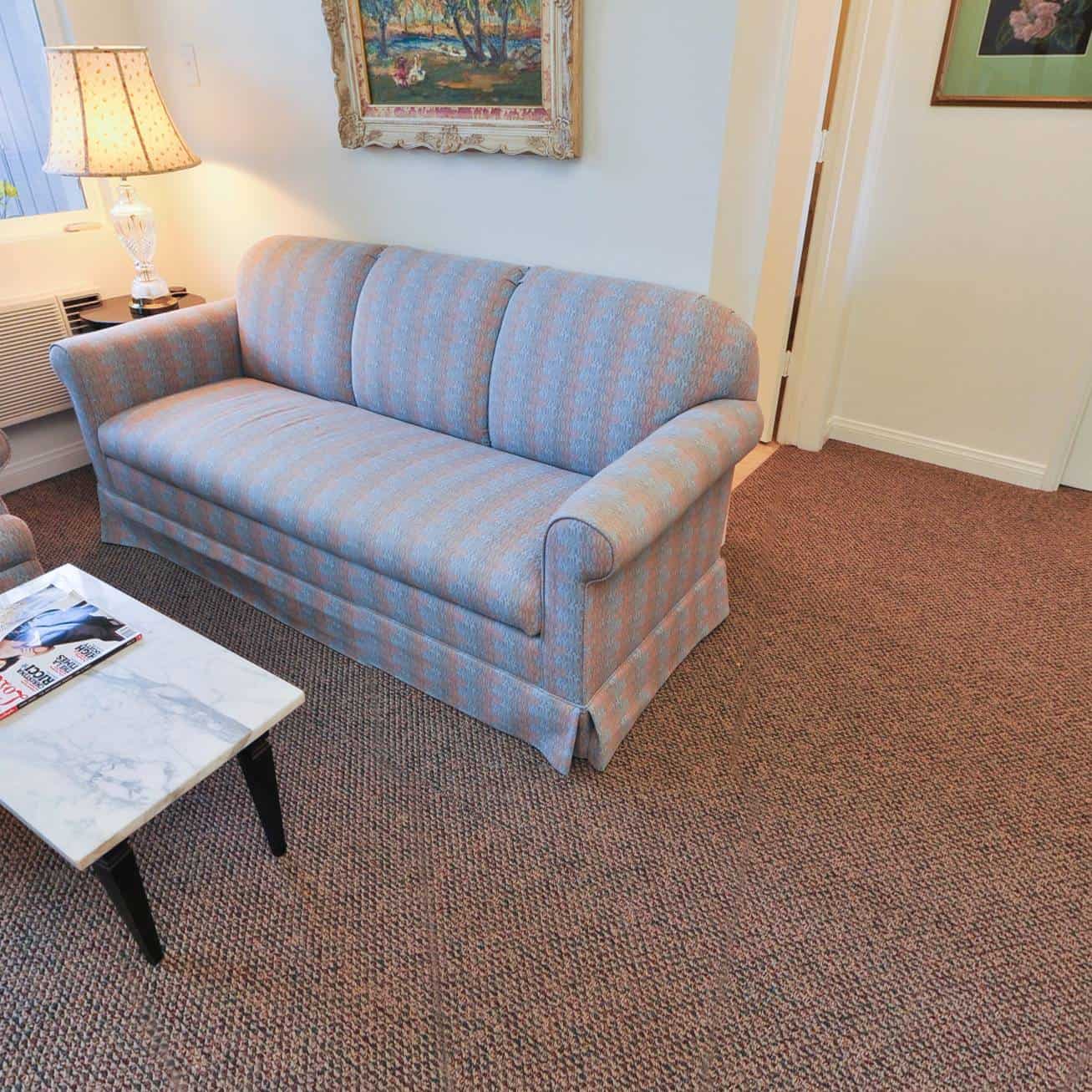 A living room featuring a blue and pink striped sofa, a marble-top coffee table with magazines, a table lamp, wall art, and carpeted floor.