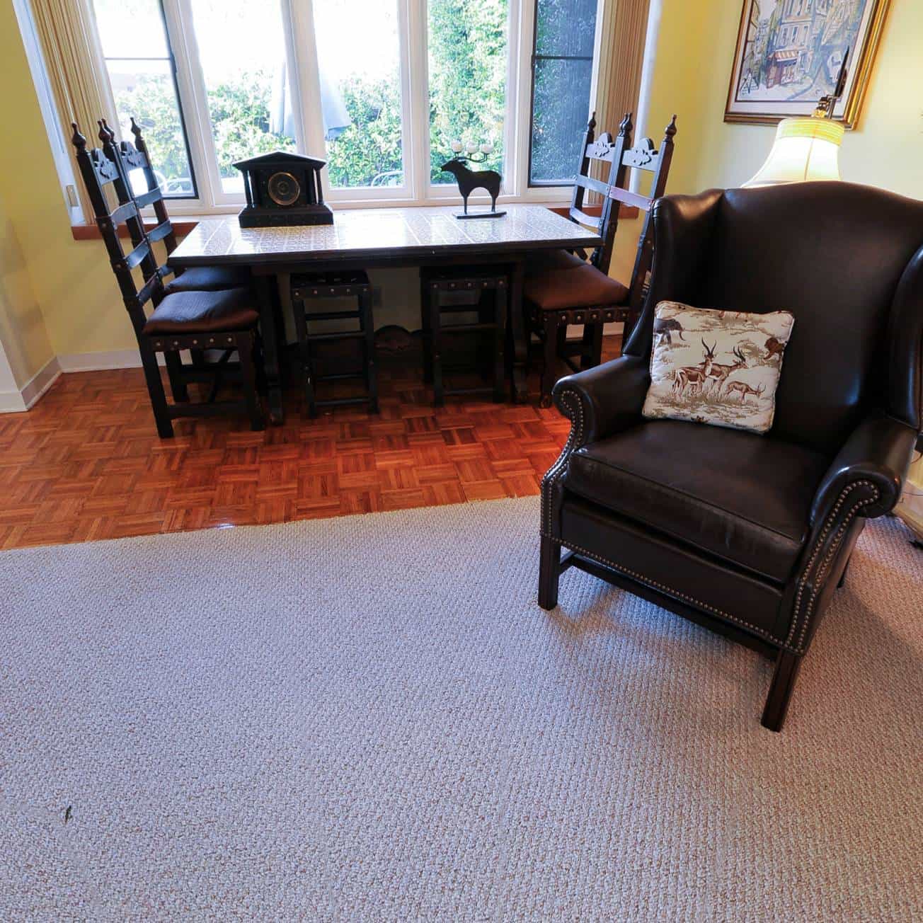 A living room with a black armchair, a dining table with black chairs, a beige carpet, and a parquet floor section by the window.