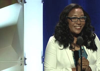 monica-macer-100th A woman in glasses is standing in front of a microphone.