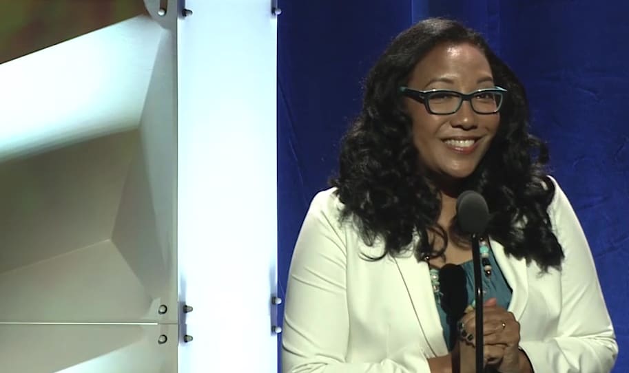 monica-macer-100th A woman in glasses is standing in front of a microphone.
