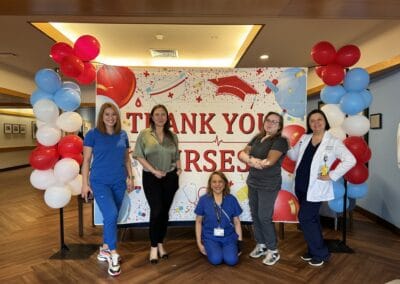 442435169_998745475146856_2670790435510543401_n Five healthcare professionals are standing in front of a "Thank You Nurses" banner decorated with balloons. Four are standing and one is kneeling, all smiling.