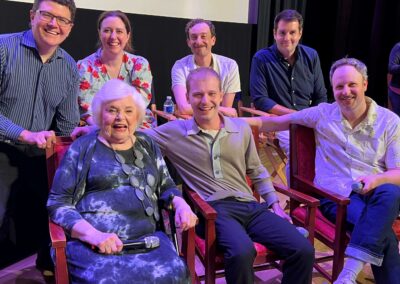 Thelma Group Q&A A group of seven people, including an elderly woman holding a microphone, are smiling and posing together in a theater-like setting. Four are seated in chairs, and three are standing behind them.